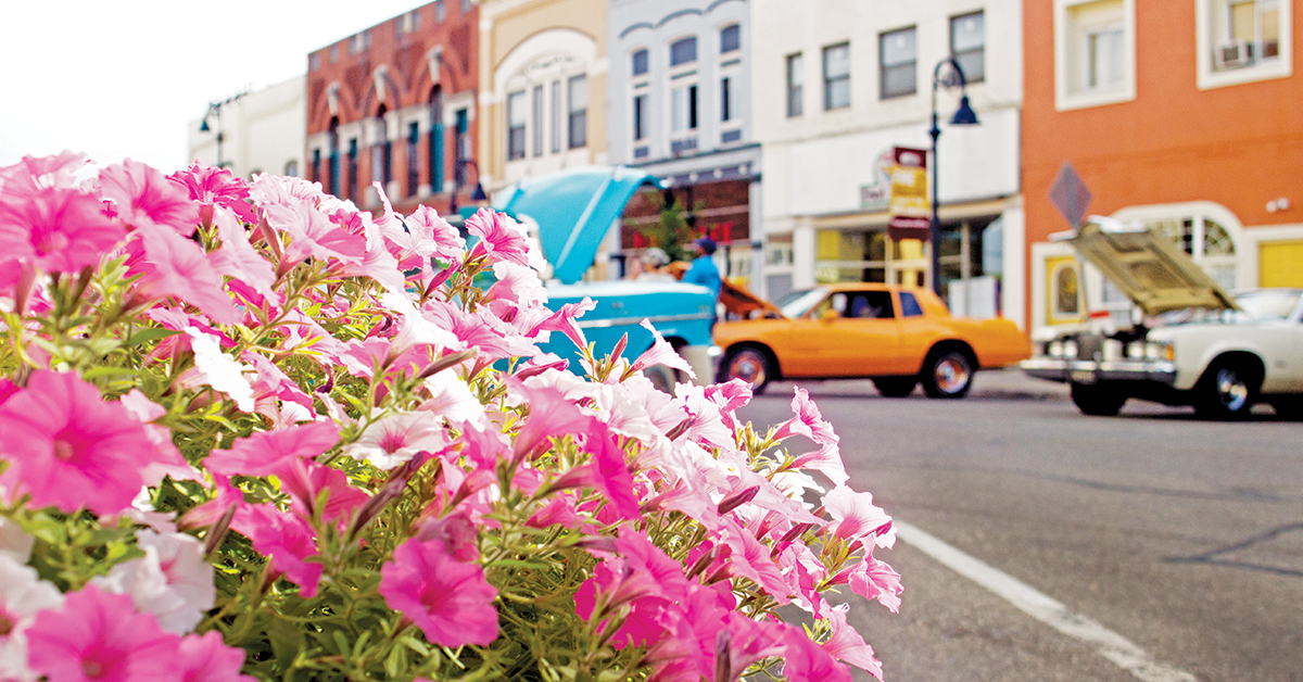 A photo of a vibrant, downtown Mt. Pleasant, Michigan street, featuring beautiful flowers in a planter and classic cars lining the streets.
